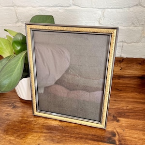 May include: A rectangular picture frame with a gold-colored textured border and a dark gray interior. The frame is set on a wooden surface, with a white brick wall in the background. A potted plant with green leaves is to the left of the frame.