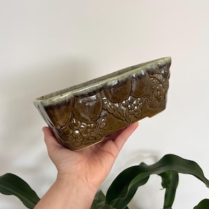 May include: A rectangular ceramic planter with a glossy olive green glaze. The exterior features a relief pattern of embossed fruit and grape clusters along the sides. The rim has a lighter, cream-colored drip glaze effect over the dark green base.