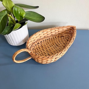 May include: A woven wicker basket with a handle. The basket is leaf-shaped and made of light brown wicker. A white potted plant with green leaves is in the background. The basket is on a blue surface.