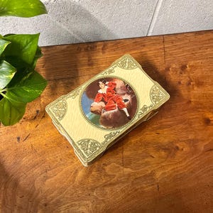 May include: A vintage rectangular trinket box with a cream-colored background and a decorative gold border. The lid features a circular image of a person in a red outfit. The box is placed on a wooden surface.