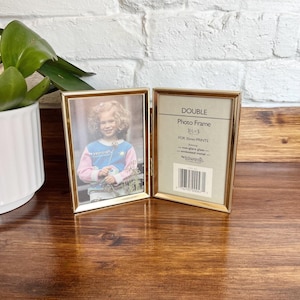 May include: A double gold-tone metal photo frame, holding a 3.5 x 5 inch photo. The frame is open, displaying a photo of a young girl. The frame's label reads "DOUBLE Photo Frame 3 1/2 x 5 FOR 35mm PRINTS".