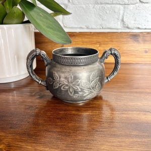 May include: An antique silver sugar bowl with two ornate handles. The bowl features a floral design on the front and is set on a wooden surface. The sugar bowl is next to a white ceramic pot with a green plant.