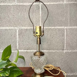 May include: A table lamp featuring a clear, diamond-patterned glass base with a brass-toned metal neck, circular base, and a wire harp. The lamp includes a gold-colored socket with a black switch and a beige electrical cord extending from the side.