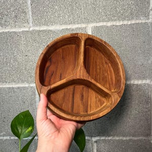 May include: A round wooden serving tray featuring three distinct, curved compartments separated by integrated dividers. The tray displays a rich, warm brown wood grain pattern with a smooth, polished finish throughout the interior and exterior.