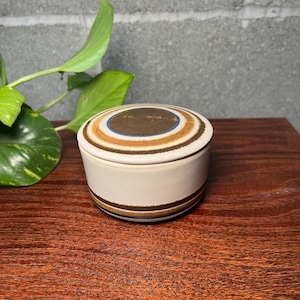 May include: A round ceramic lidded container featuring a cream-colored base with horizontal brown and blue striped bands. The lid displays a concentric circular pattern in shades of brown, tan, and blue, centered on a dark brown circular disc.