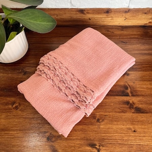 May include: Folded pink scarf with a decorative lace trim. The scarf is made of a textured fabric and is neatly folded. The scarf is displayed on a wooden surface, with a white pot and green plant in the background.
