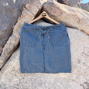 May include: Blue and tan plaid skirt hanging on a wooden hanger. The skirt has a drawstring waist and two front pockets. The fabric has a grid pattern. The skirt is displayed against a backdrop of large rocks.