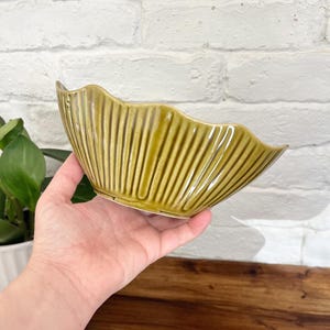 May include: A vintage, olive-green ceramic bowl with a scalloped rim and vertical ribbed detailing. The bowl is held in a hand, showcasing its shape and texture. The background includes a white brick wall and a wooden surface.