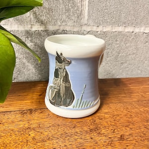 May include: A ceramic mug with a light blue exterior and a white rim. The mug features a black and gray illustration of a golf bag. The mug is sitting on a wooden surface.