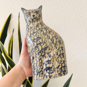 May include: A ceramic cat figurine with a blue and white speckled glaze. The cat is sitting with its head tilted slightly to the side.