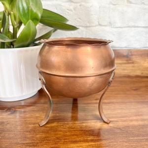 May include: A copper-colored bowl with a scalloped rim, supported by three curved legs. The bowl has a rounded shape and a polished finish. A white ceramic pot with a green plant is in the background.