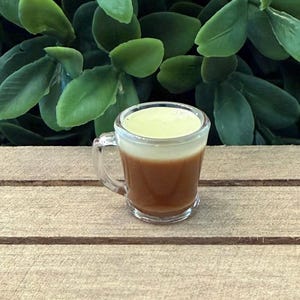 May include: A small clear glass mug filled with a layered beverage. The bottom layer is brown, the middle layer is a lighter brown, and the top layer is a pale yellow. The mug has a handle and sits on a wooden surface.