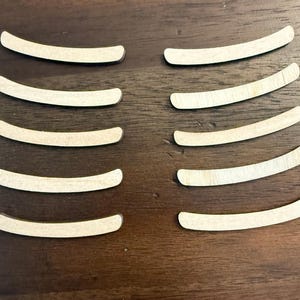 May include: Ten unfinished, curved wooden pieces arranged on a dark brown wooden surface. Each piece is a light tan color, with a smooth, slightly rounded shape. The pieces are evenly spaced.
