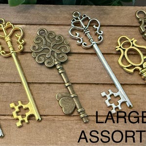 May include: Assorted large decorative keys in silver, gold, and bronze tones. The keys have ornate designs, including filigree and floral patterns. The keys are arranged on a wooden surface. Text on the image reads "LARGE ASSORTED".