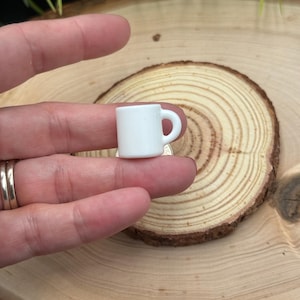May include: A small, white ceramic mug with a handle. The miniature mug is held in a person's hand and is set against a natural wood slice background. The mug is a simple, classic design.