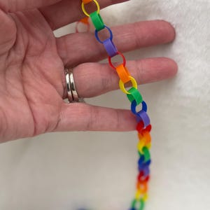 May include: A colorful paper chain in a rainbow of hues, including red, orange, yellow, green, blue, and purple. The chain is held in a hand against a soft, white background.