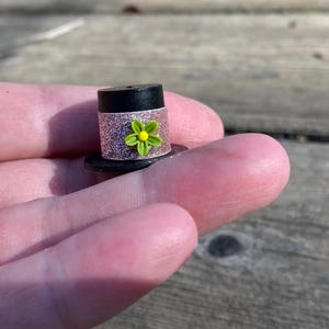 May include: A miniature black top hat with a pink glitter band and a green flower with a yellow center.