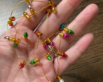 May include: A string of miniature Christmas lights with colorful bulbs in red, green, pink, blue, and silver. The lights are strung on a thin gold wire. The lights are held in a hand.