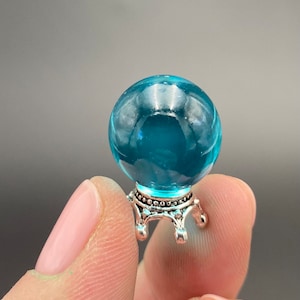 May include: A small, teal blue glass orb sits on a silver stand with three legs. The orb is clear and reflects light.