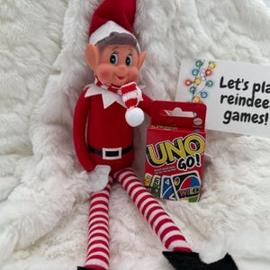 May include: A red and white elf doll wearing a matching hat and scarf, with a black belt and striped legs. The elf is holding a red box of UNO GO! cards and a sign that reads "Let's play reindeer games!"