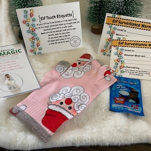 May include: A pink elf glove with a Santa Claus design, a small bottle of glitter, and two Elf Surveillance Warning cards with a yellow and black striped border. The cards have blank spaces for the elf's name and the behavior observed. The text on the cards reads "Elf Surveillance Warning".