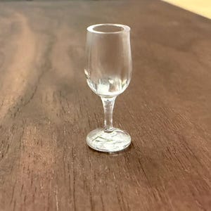 May include: A small, clear glass wine tasting glass with a rounded bowl and a slender stem. The glass sits on a round base. The glass is on a dark brown wooden surface.