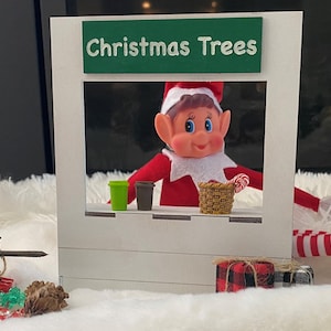 May include: A Christmas elf doll wearing a red and white striped outfit is peeking out of a wooden window frame. The window frame has a green sign that says "Christmas Trees".