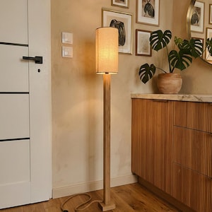 Puede incluir: Una lámpara de pie de madera con una pantalla rectangular de lino. La lámpara tiene un acabado de madera natural y se apoya sobre una base cuadrada. La lámpara está en una habitación con un mueble de madera y obras de arte enmarcadas. La lámpara mide unos 150 cm de altura.