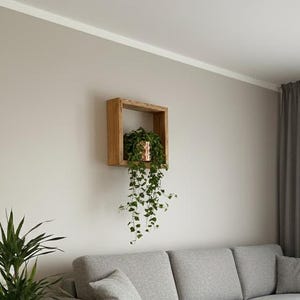 Op de afbeelding: Een moderne woonkamer met een lichtgrijze hoekbank, een ronde houten salontafel en een patroonkleed. Een potplant en een hangplant voegen een vleugje natuur toe. Grijze gordijnen en een houten plank maken de inrichting compleet. De bank is ongeveer 250 cm lang.