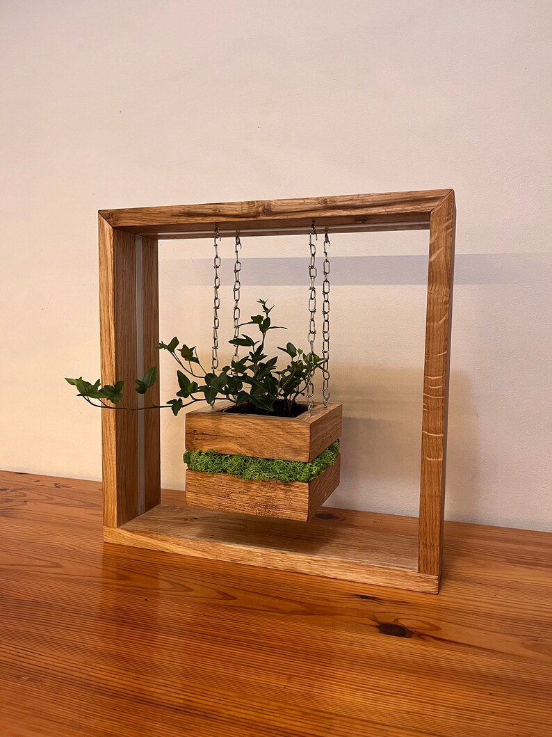 May include: A wooden hanging planter with a square frame. The planter box, filled with greenery and moss, is suspended by chains. The frame and planter are made of light-colored wood, creating a natural aesthetic. The design is suitable for indoor or outdoor use.