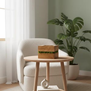 May include: A wooden box with a layer of green moss sits on a round, light wood side table. A small white vase and two books are on the lower shelf. A white armchair and a large potted plant are in the background.