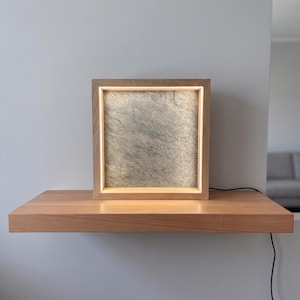 Può includere: Una lampada quadrata con cornice in legno dotata di un pannello centrale testurizzato grigio simile alla pietra, illuminato da una fonte luminosa interna. La lampada è realizzata in legno chiaro e include un sottile cavo nero sul lato.