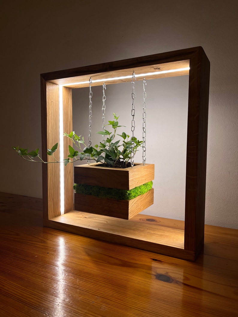 May include: A wooden planter box with greenery, suspended by chains within a rectangular wooden frame. The frame has a built-in LED light strip. The planter box is filled with soil and green plants, with a layer of green moss.