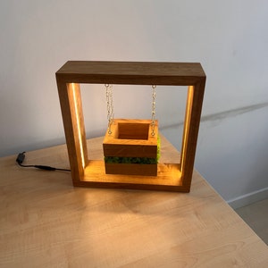 May include: A wooden lamp with a square frame and a suspended planter box. The planter is filled with green moss and hangs from gold chains. The frame has a warm light strip. The lamp is on a light wood surface.
