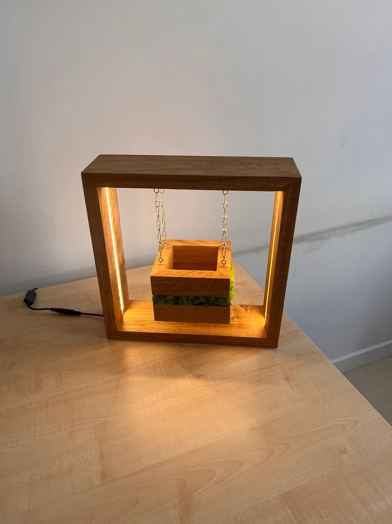 May include: A wooden lamp with a square frame and a suspended planter box. The planter is filled with green moss and hangs from gold chains. The frame has a warm light strip. The lamp is on a light wood surface.