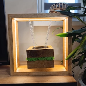 May include: A wooden planter box suspended by silver chains within a square, illuminated wooden frame. The planter is filled with soil and green moss. The frame has a warm, glowing light.