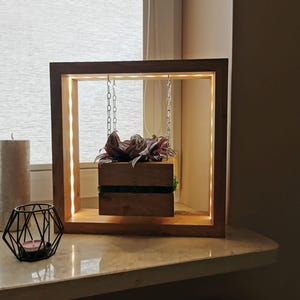 May include: A wooden planter box suspended within a square wooden frame with integrated LED lighting. The planter, filled with greenery, hangs by chains. A decorative candle holder and a pillar candle are also visible.