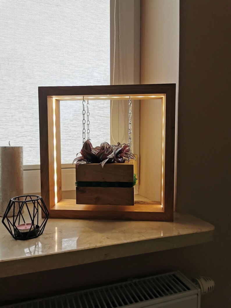 May include: A wooden planter box suspended within a square wooden frame with integrated LED lighting. The planter, filled with greenery, hangs by chains. A decorative candle holder and a pillar candle are also visible.