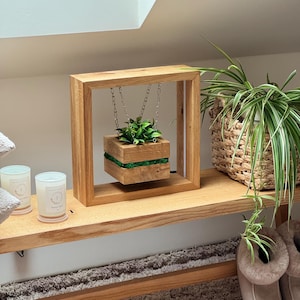 Puede incluir: Una jardinera flotante de madera con un marco cuadrado y una jardinera suspendida. La jardinera está llena de vegetación y acentuada con una franja verde. El marco es de madera clara.