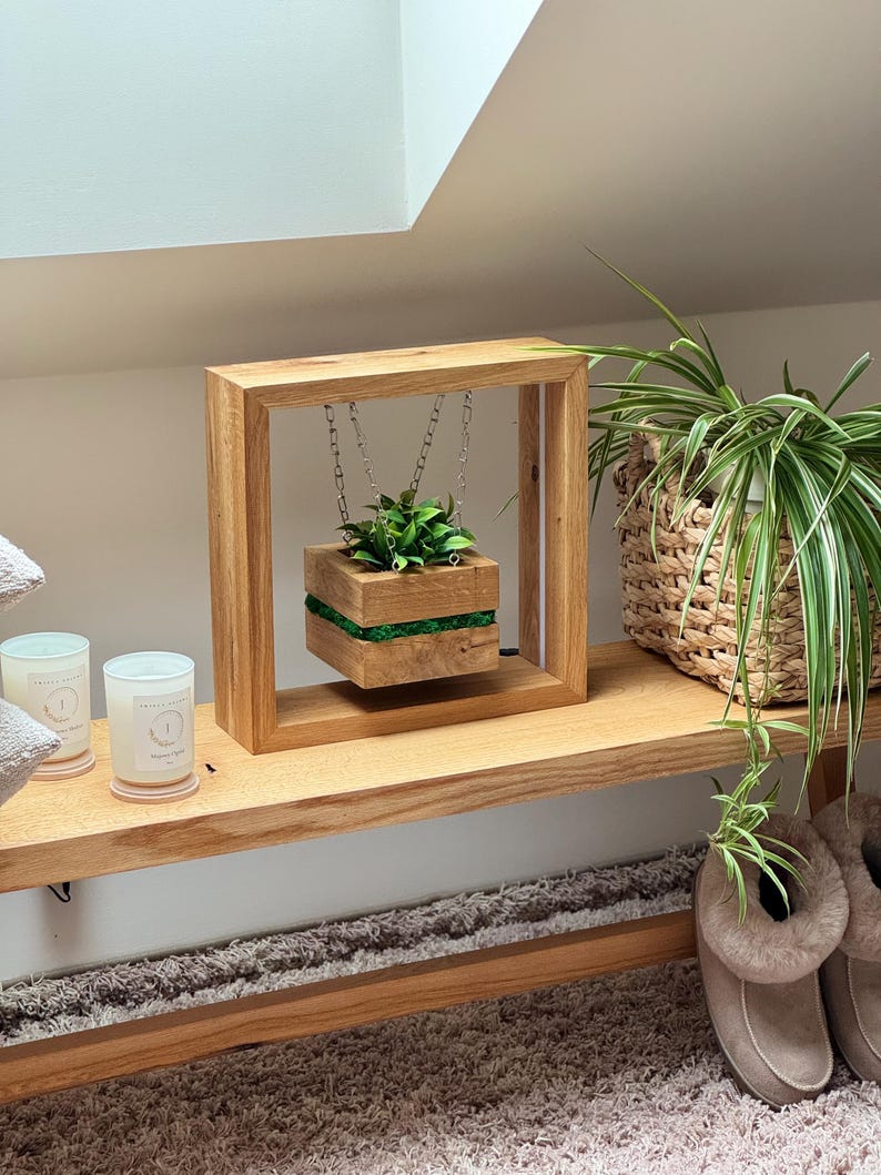 May include: A wooden floating planter with a square frame and a suspended planter box. The planter box is filled with greenery and accented with a green stripe. The frame is made of light-colored wood.