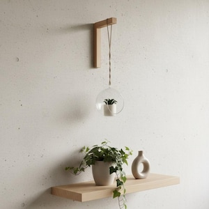Op de afbeelding: Een minimalistisch interieur met een houten plank en een hangend glazen terrarium. Een kleine plant staat in het terrarium, opgehangen aan een bruin koord. Een witte vaas en een potplant met groene bladeren staan op de plank.