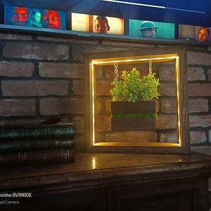 May include: A wooden framed wall art piece with a square shape, featuring a small plant in a dark wooden planter. The frame has a warm, yellow LED light. The art piece hangs in front of a brick wall.