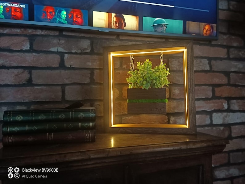 May include: A wooden framed wall art piece with a square shape, featuring a small plant in a dark wooden planter. The frame has a warm, yellow LED light. The art piece hangs in front of a brick wall.