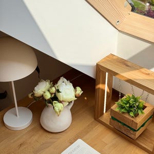 May include: A serene interior scene featuring a white table lamp, a small white vase with white flowers, and an open book with the text "RYTM NATURY". A wooden planter with greenery hangs from a wooden frame.