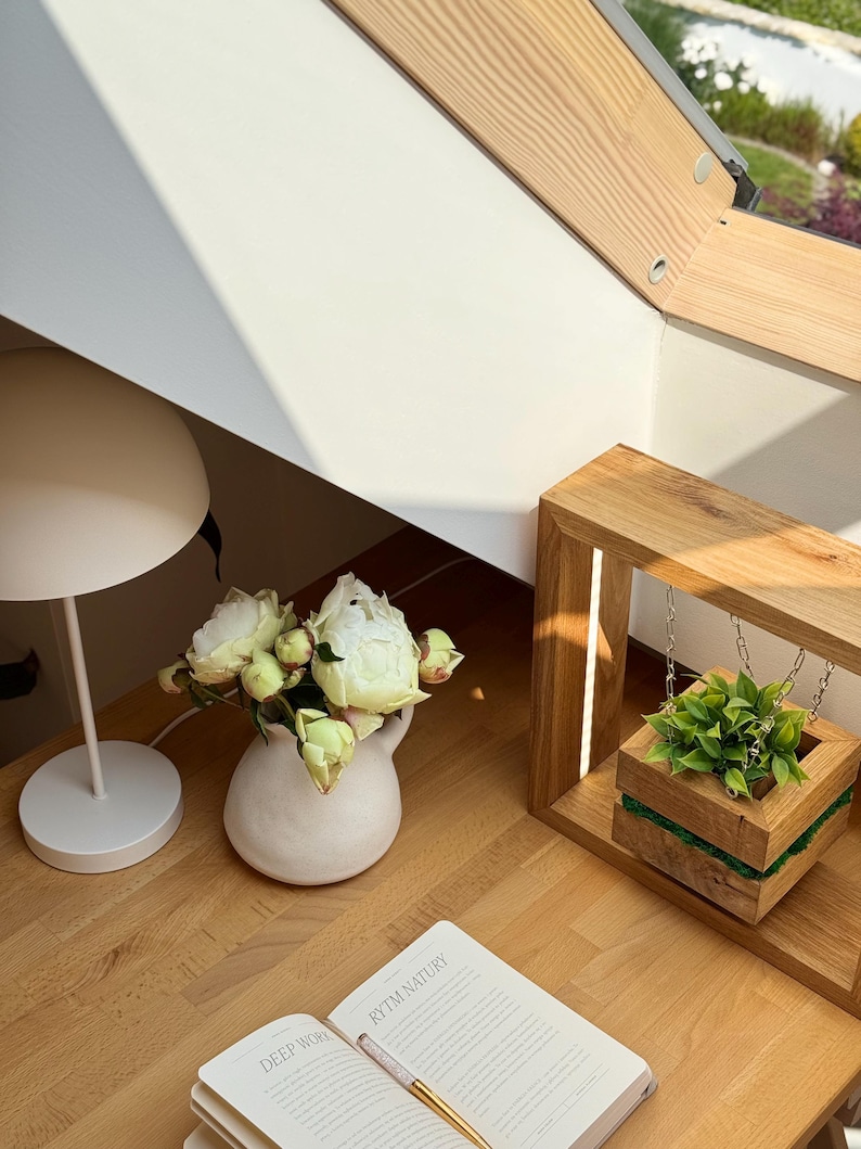 May include: A serene interior scene featuring a white table lamp, a small white vase with white flowers, and an open book with the text "RYTM NATURY". A wooden planter with greenery hangs from a wooden frame.