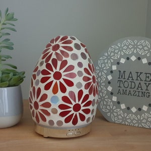 May include: A white and red mosaic diffuser with a gold base and two buttons. The diffuser is shaped like an egg and has a floral pattern. There is a small potted succulent plant and a grey sign with the text "MAKE TODAY AMAZING" in the background.