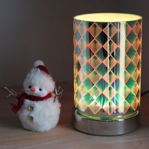 May include: A silver and glass electric wax warmer with a geometric pattern in shades of green, orange, and yellow. The warmer is illuminated from within, casting a warm glow.