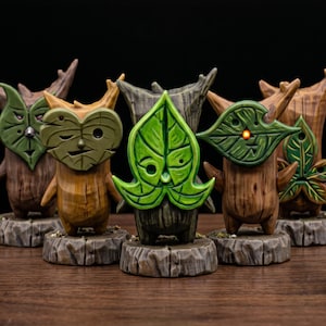 Peut inclure: Une collection de cinq petites figurines à la texture de bois, debout sur des socles circulaires. Chaque personnage porte un masque sculpté en forme de feuille couvrant son visage, dont l'un présente une petite lumière orange brillante.