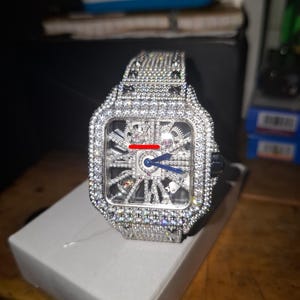 May include: A luxury wristwatch with a square face and a silver band. The watch is encrusted with numerous small, sparkling stones. The watch face has a complex, open design, revealing the inner workings. The hands are blue.