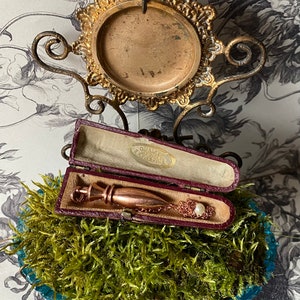 Op de afbeelding: Een goudkleurige, traanvormige hanger met een parel accent, rustend in een bordeauxrode fluwelen doos. De doos is open en de hanger is zichtbaar. De hanger zit aan een delicate ketting.